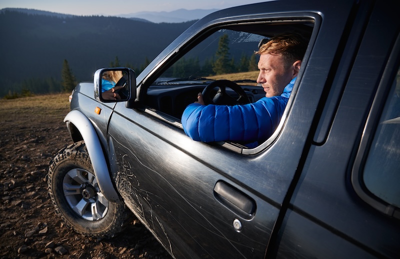 Close up view of young male blonde driving black SUV, looking at landscape from car, and riding on dirt roads in the mountains. Extreme off-road riding on mountain hills.