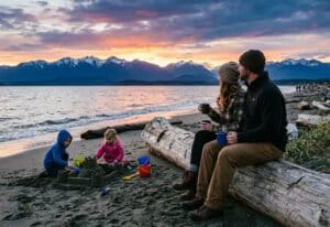 two people sitting on a log on a beach in Victoria at sunset while their children play in the sand near the water