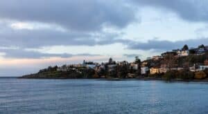 A serene morning scene in Victoria, Vancouver Island, BC, showcasing charming coastal homes under the soft light of sunrise with a tranquil sea view. The calm sky complements the picturesque landscape.