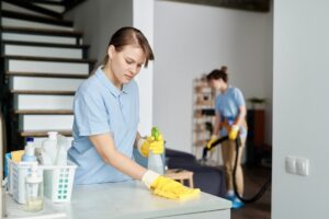 Cleaning service worker wiping dust from furniture with rag and detergent with her colleague vacuuming in background