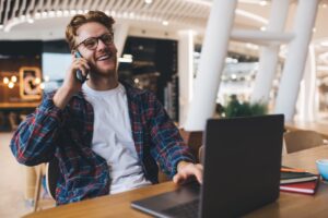 Young caucasian guy talking on smartphone at table with opened laptop. Concept of online education and job. Idea of modern successful man. Smiling guy in glasses working or studying at office space 