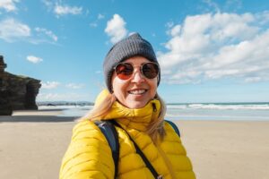 Young woman smiling on camera with beach in background during winter time - Travel destination concept - Focus on face