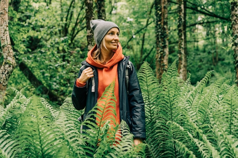 Young, blonde woman wearing light layers and a touque in the forest surrounded by ferms - pacific northwest fashion, packing, what to wear